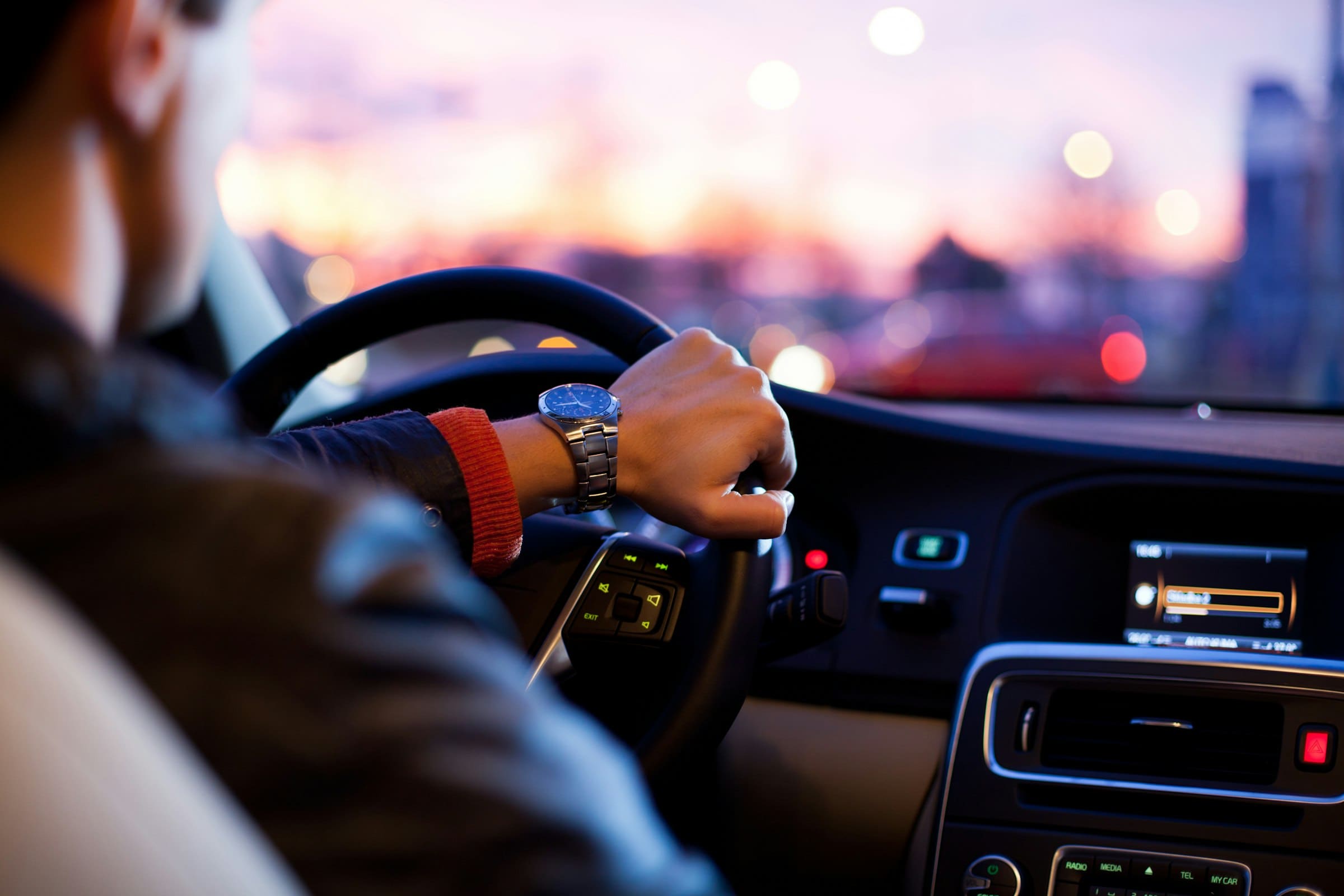 A driver's hands on the steering wheel of a car at dusk, city lights beyond the windscreen