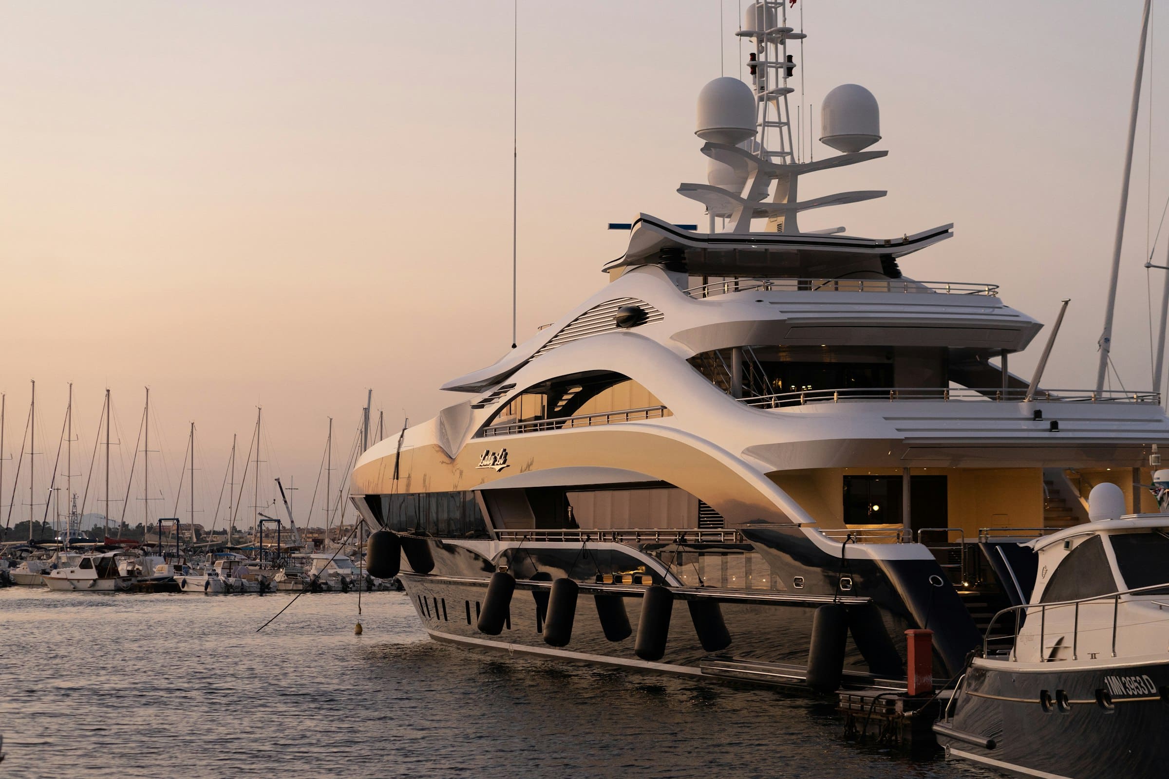 A superyacht moored in a Mediterranean marina at golden hour, its decks lit by the evening sun