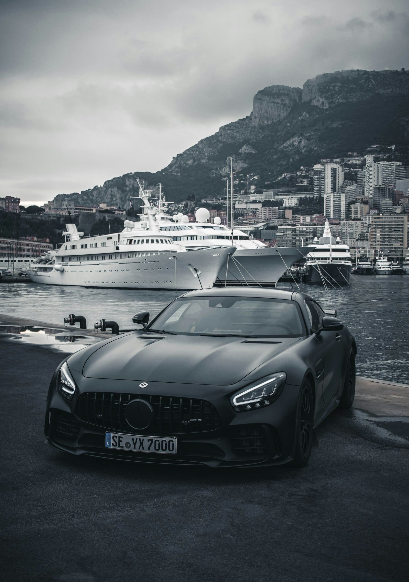 A dark luxury car parked on a harbour front with yachts and a hillside town behind